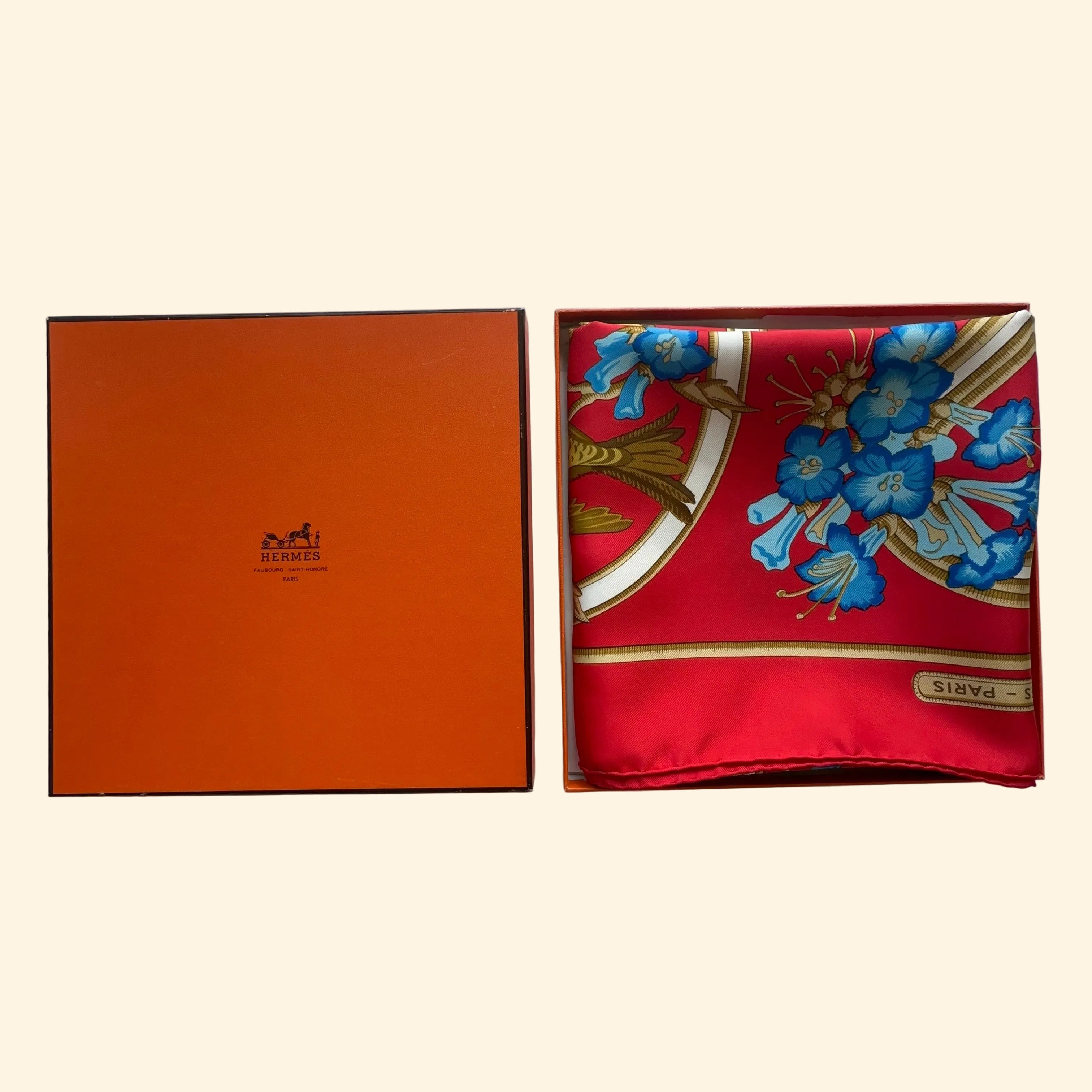 Le carré Hermès Caraïbes rouge soigneusement plié à l'intérieur de sa boîte orange d'origine, révélant ses motifs de fleurs bleues.