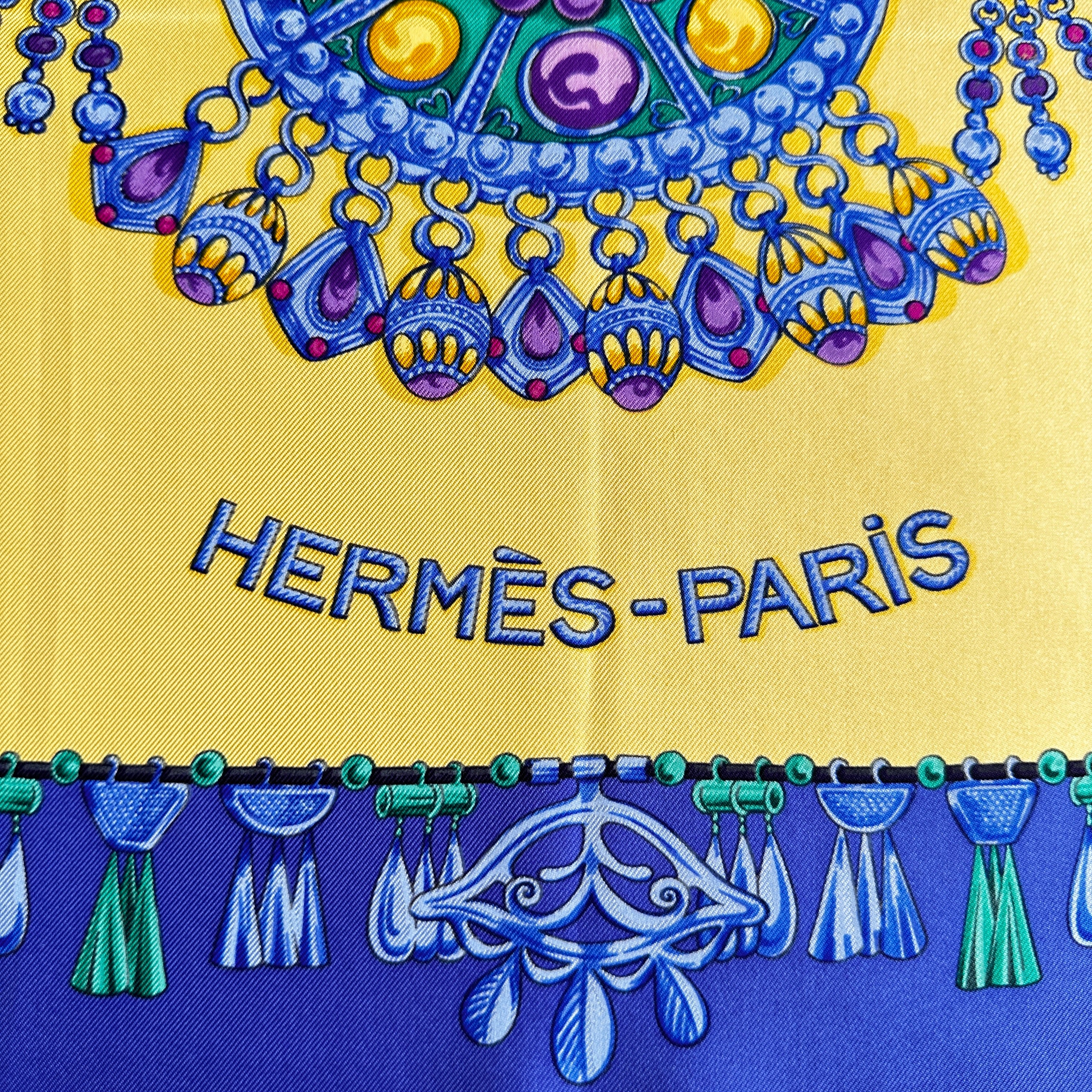 Inscription Hermès-Paris et détail des franges de bijoux bleus sur fond jaune.