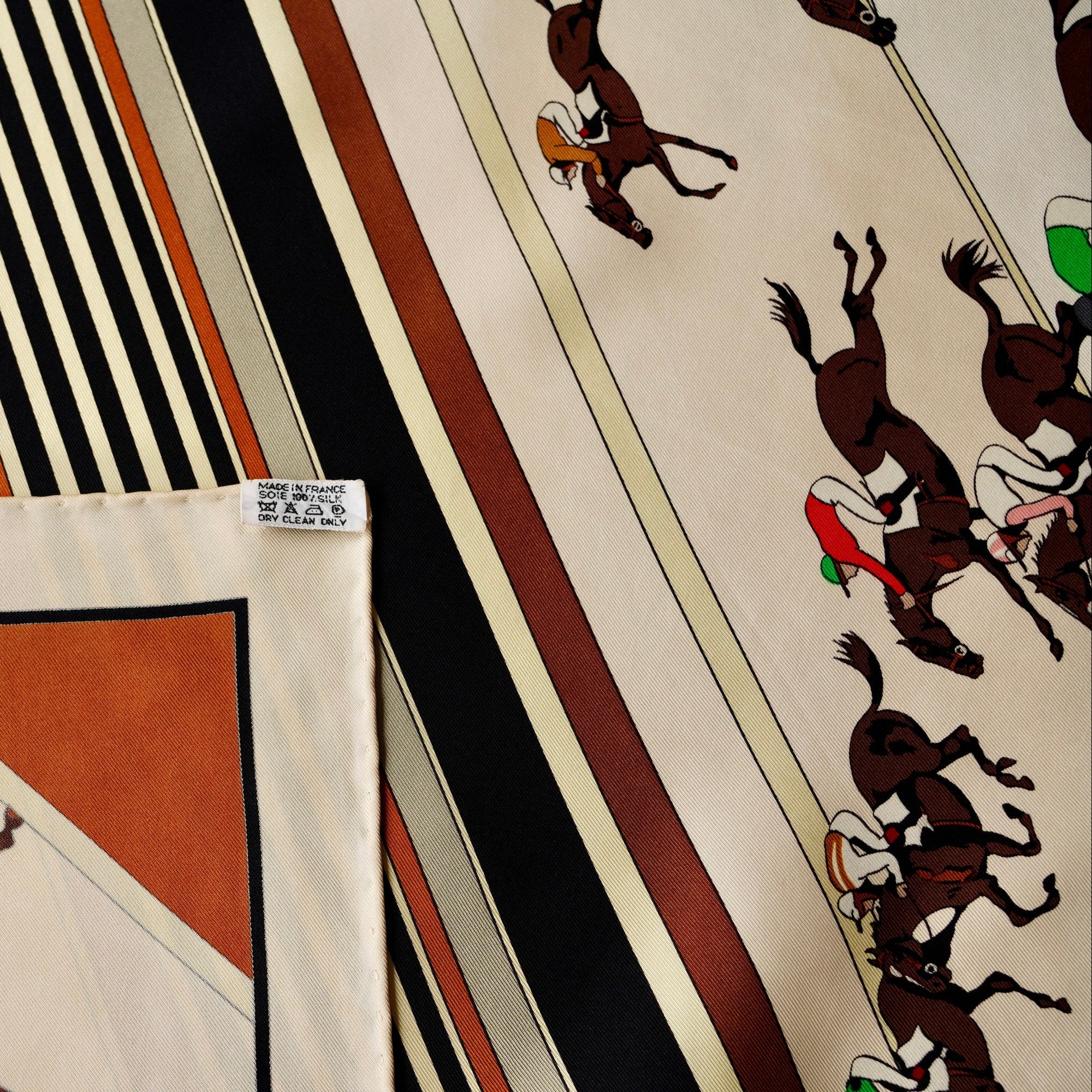 Zoom sur une petite tache discrète sur le fond crème du foulard Hermès "Les Courses", pointée par deux flèches bleues.